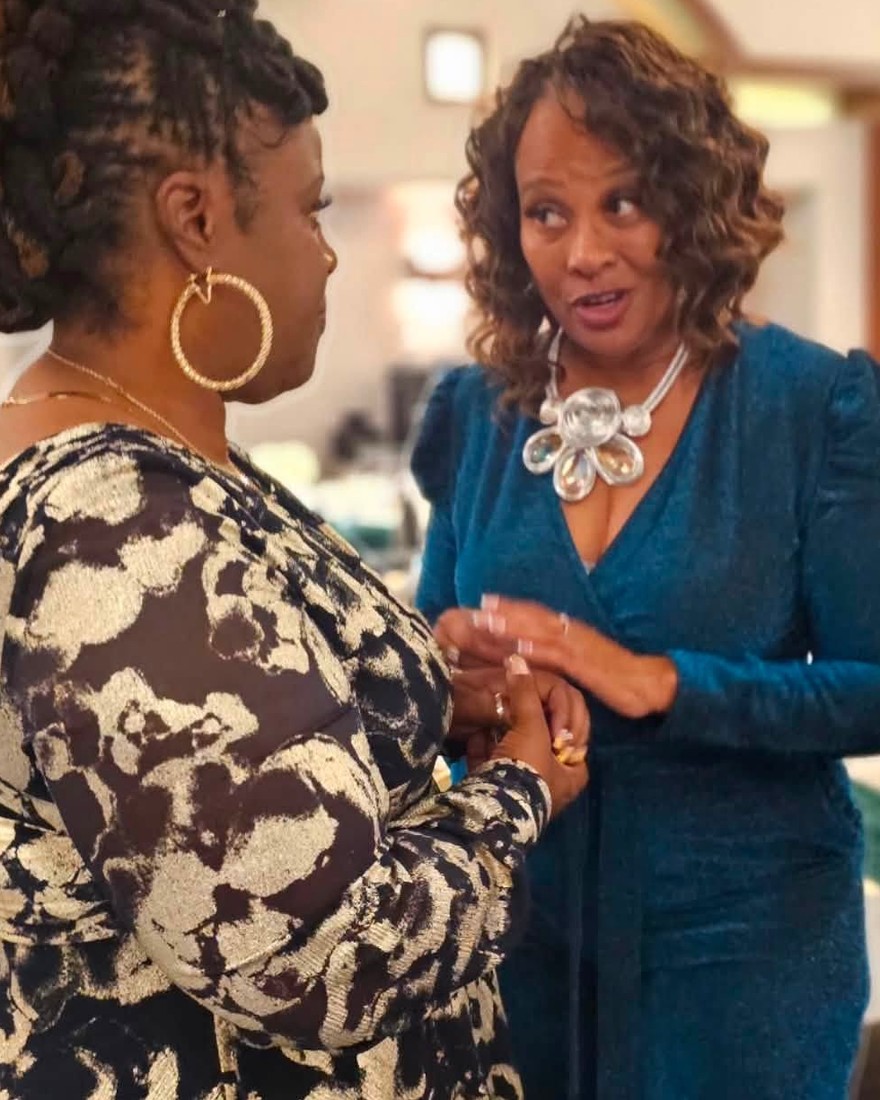 Carole Pinkney-Hart in a teal ensemble, sharing a warm, attentive moment with a woman at an event